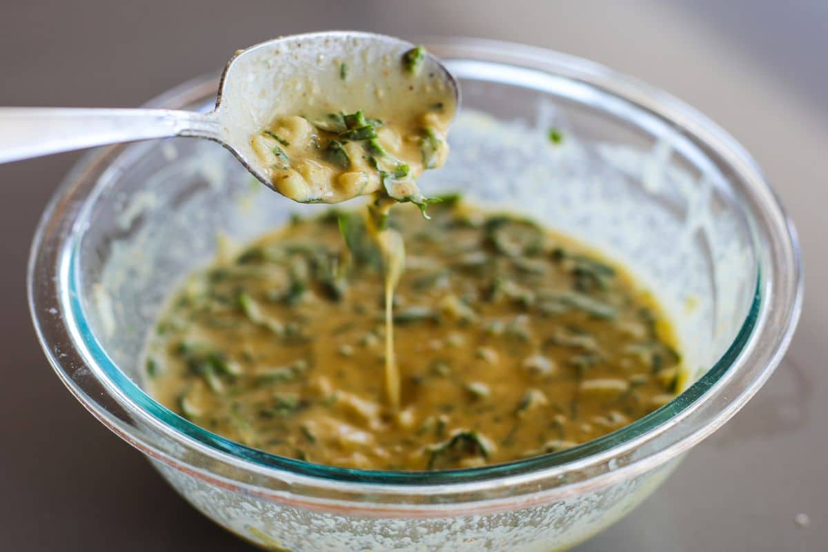 A metal spoon holds up a flowy besan batter with veggies.