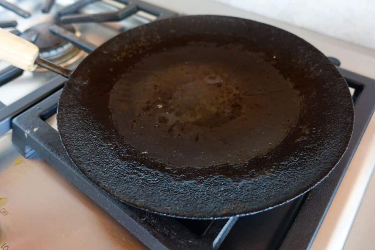 A round, black, cast iron griddle sits on a stovetop burner, showing signs of use with oil and seasoning on its surface.
