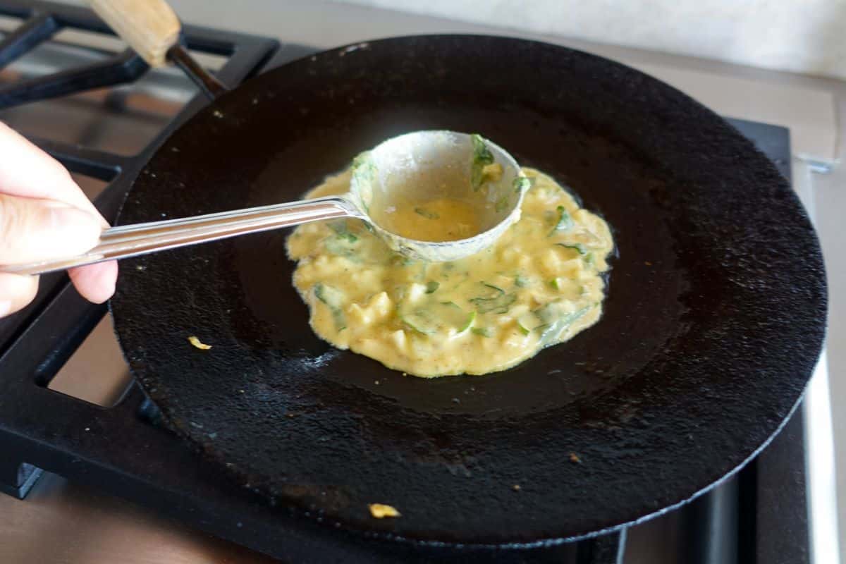 A hand pours a ladle of batter with chopped greens onto a black pan on a stovetop for cooking.