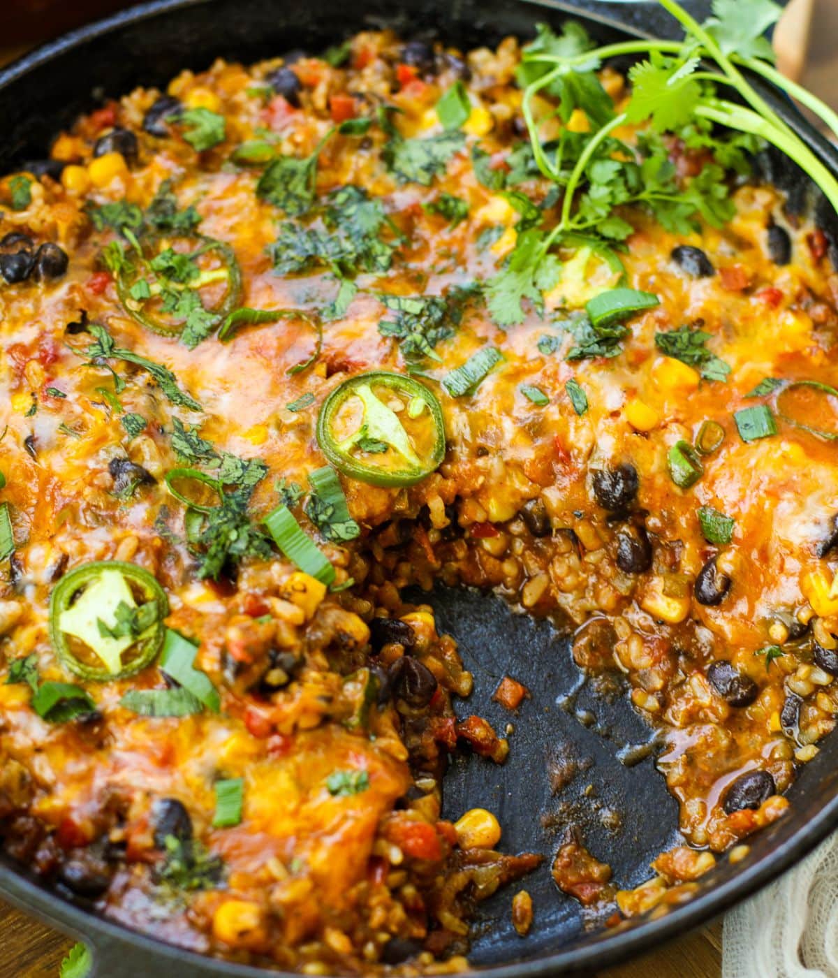 An easy vegetarian burrito skillet of cheesy Mexican rice with black beans, corn, bell peppers, and jalapeño slices, garnished with cilantro. A serving has been removed, revealing the inside.