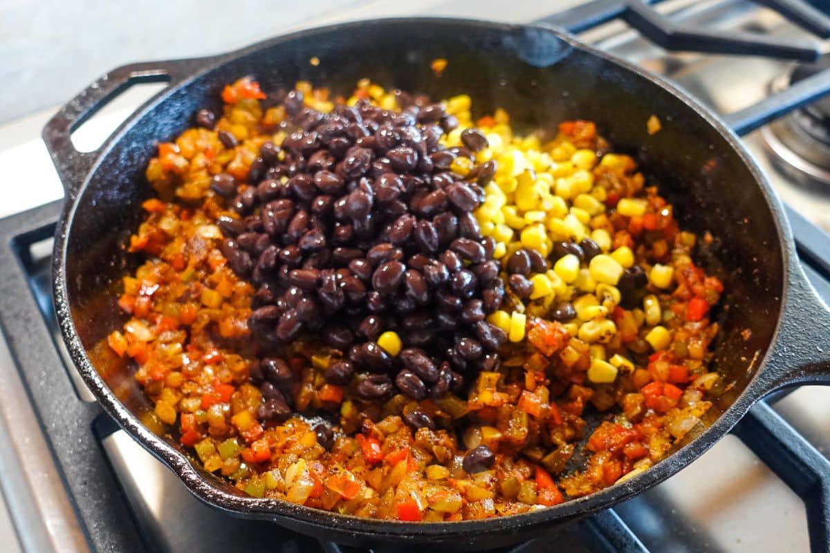 A cast iron skillet on a stove containing sautéed diced vegetables, topped with black beans and corn.