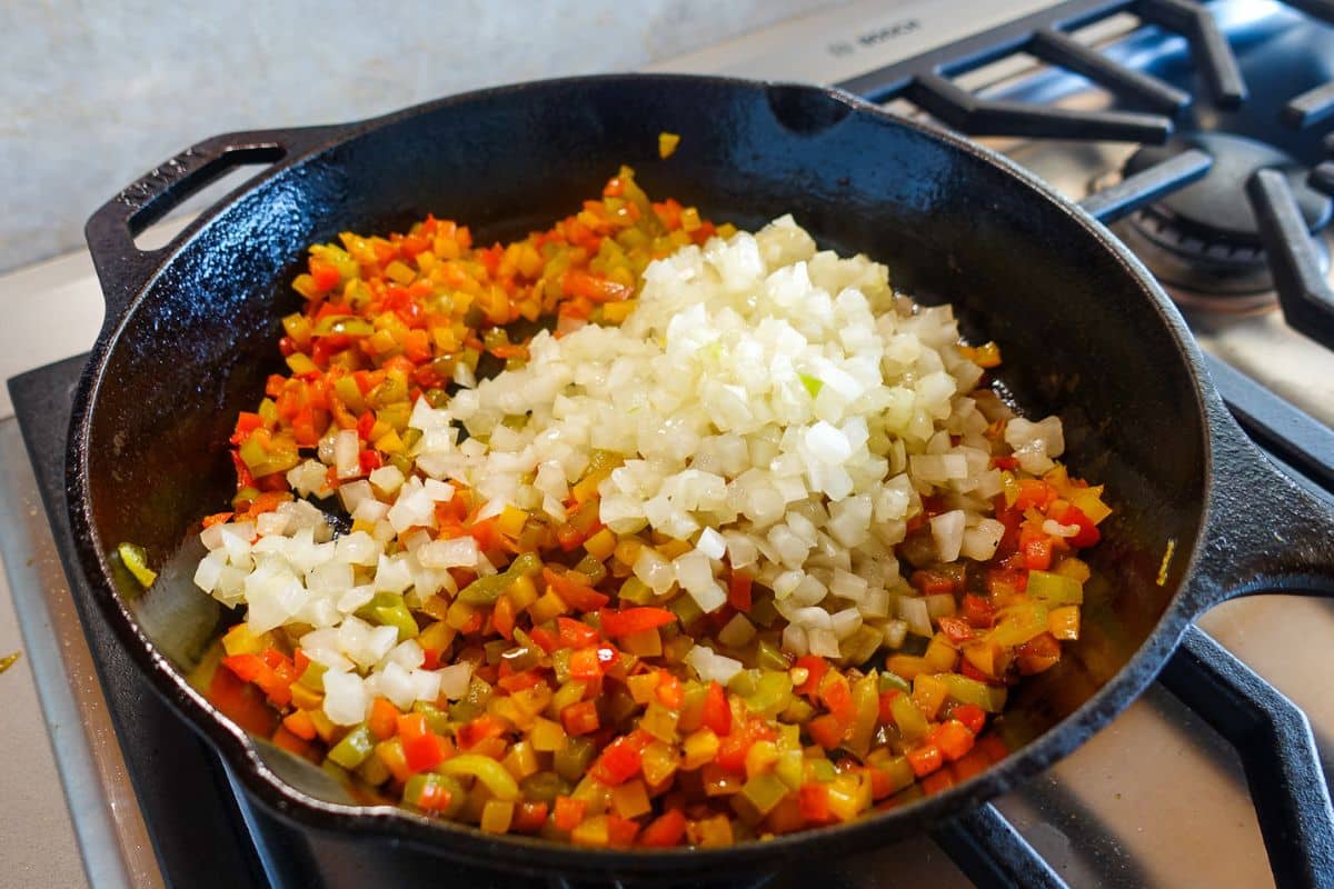 Diced red and green bell peppers and chopped onions cooking in a cast iron skillet on a gas stove.