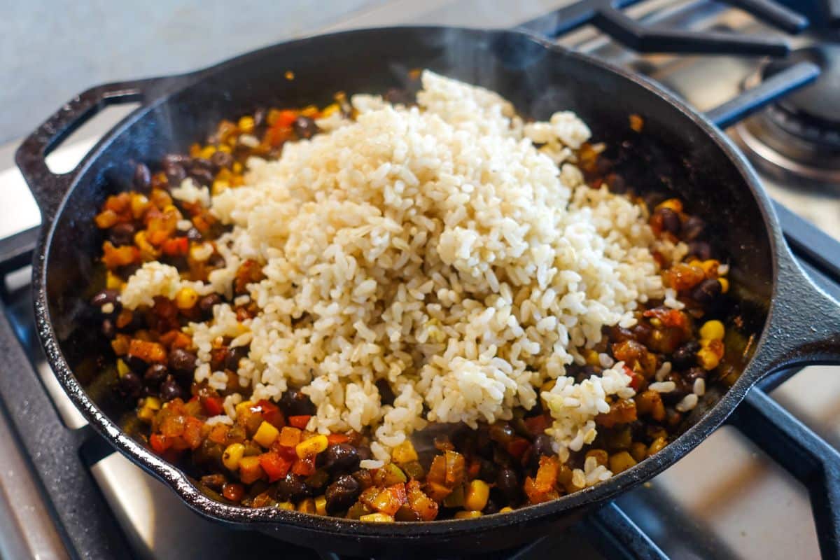 A cast iron skillet on a stove contains sautéed vegetables, black beans, corn, and a mound of cooked brown rice on top.