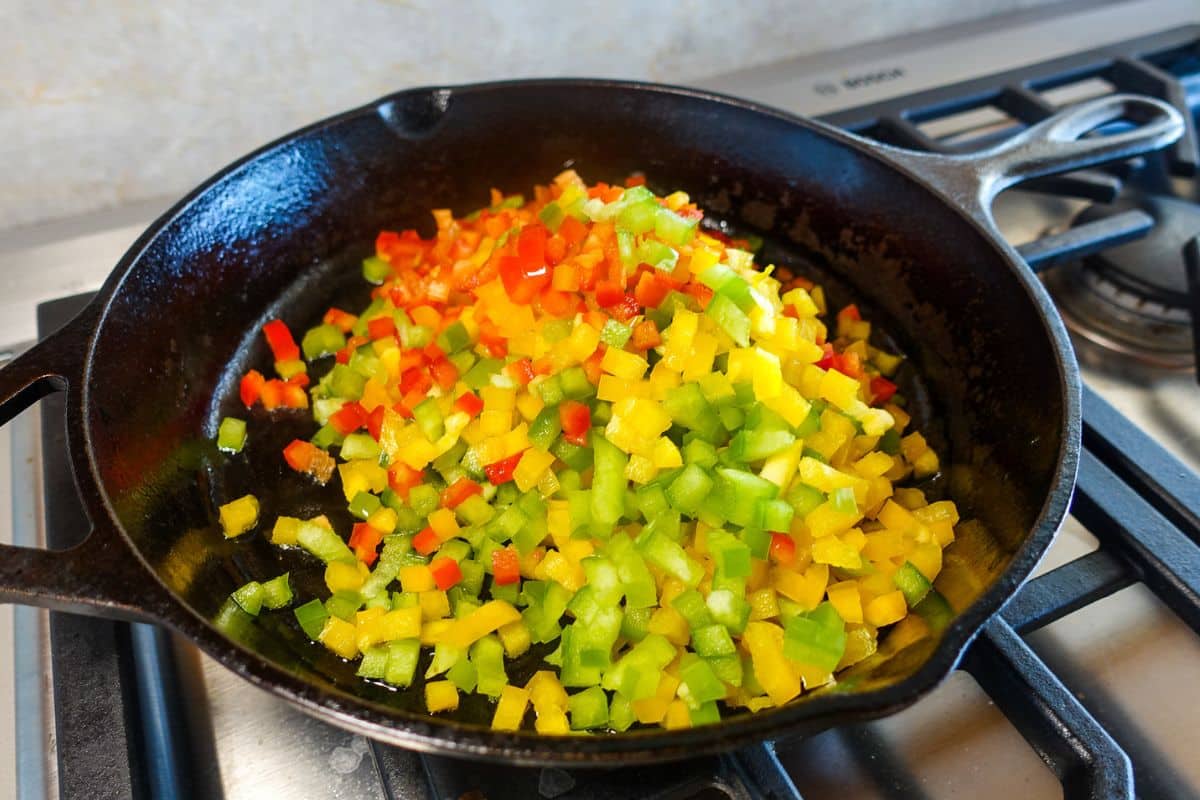 Diced red, yellow, and green bell peppers in a black cast iron skillet on a stovetop.