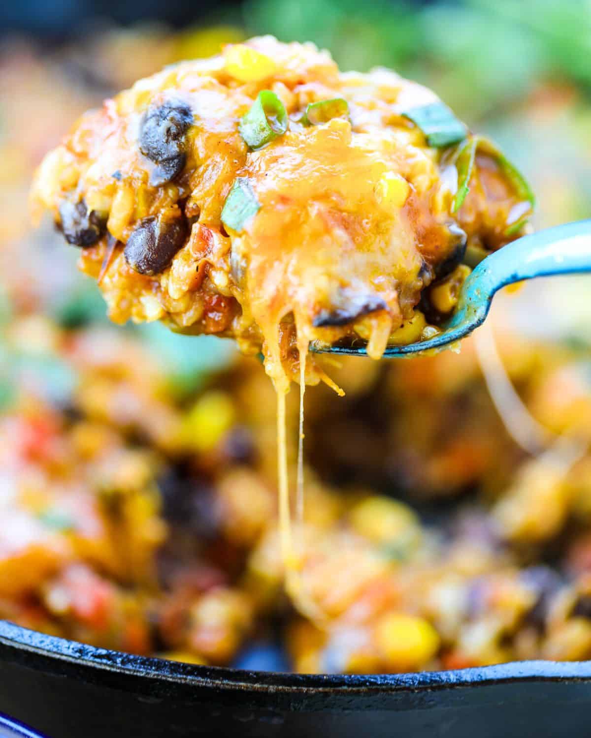 Vegetarian Burrito Skillet close-up of a spoon lifting a cheesy, baked mixture with black beans, corn, and chopped vegetables from a skillet.