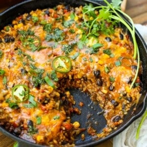 A Vegetarian Burrito Skillet filled with a cheesy baked casserole containing black beans, corn, rice, vegetables, and sliced jalapeños, garnished with fresh cilantro, with a serving missing.