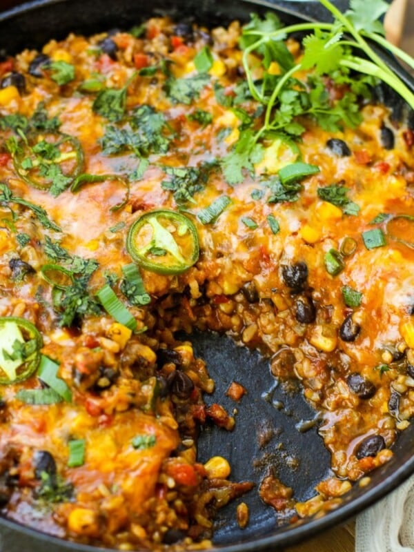 A Vegetarian Burrito Skillet filled with a cheesy baked casserole containing black beans, corn, rice, vegetables, and sliced jalapeños, garnished with fresh cilantro, with a serving missing.