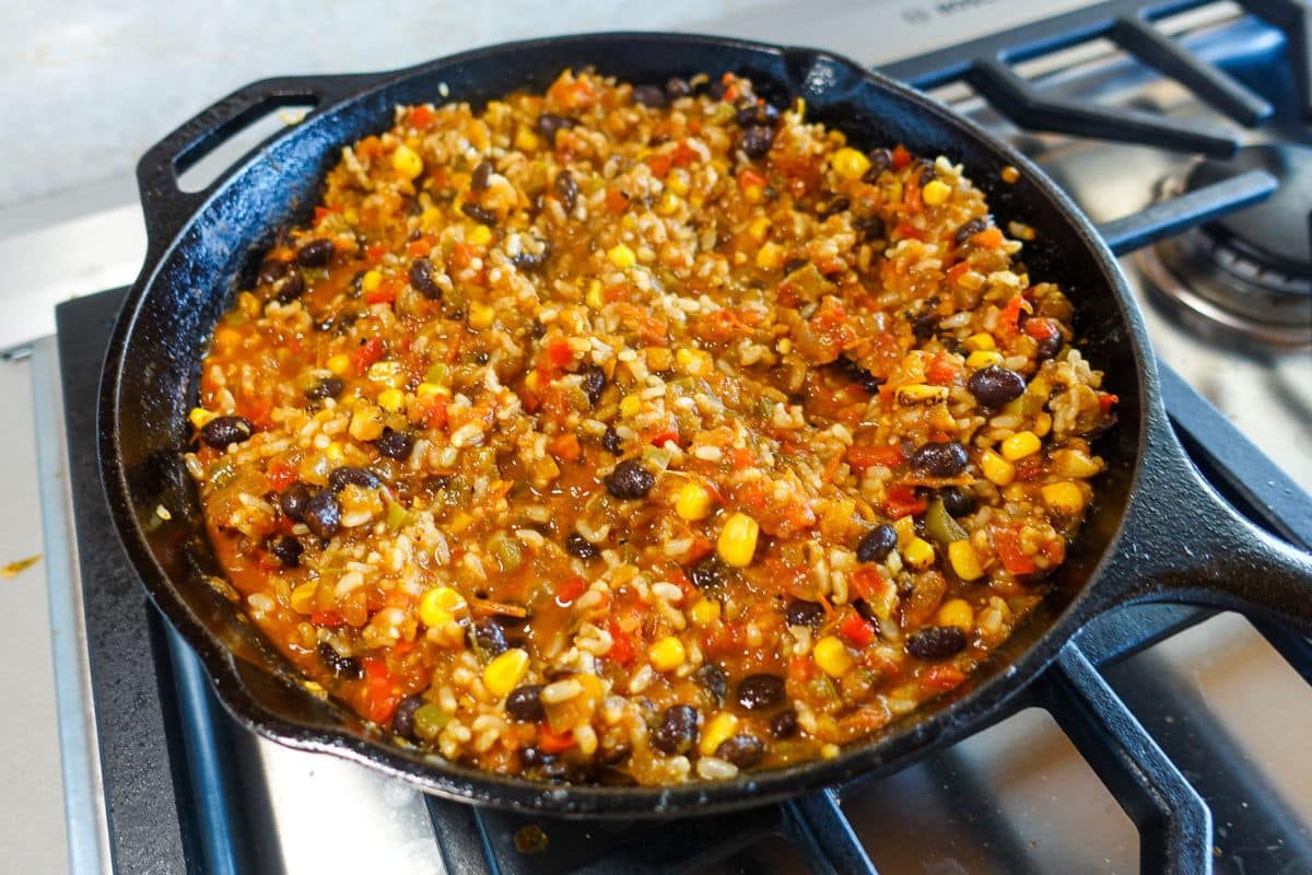 A skillet on a stove contains a cooked mixture of rice, black beans, corn, salsa, and diced bell peppers.
