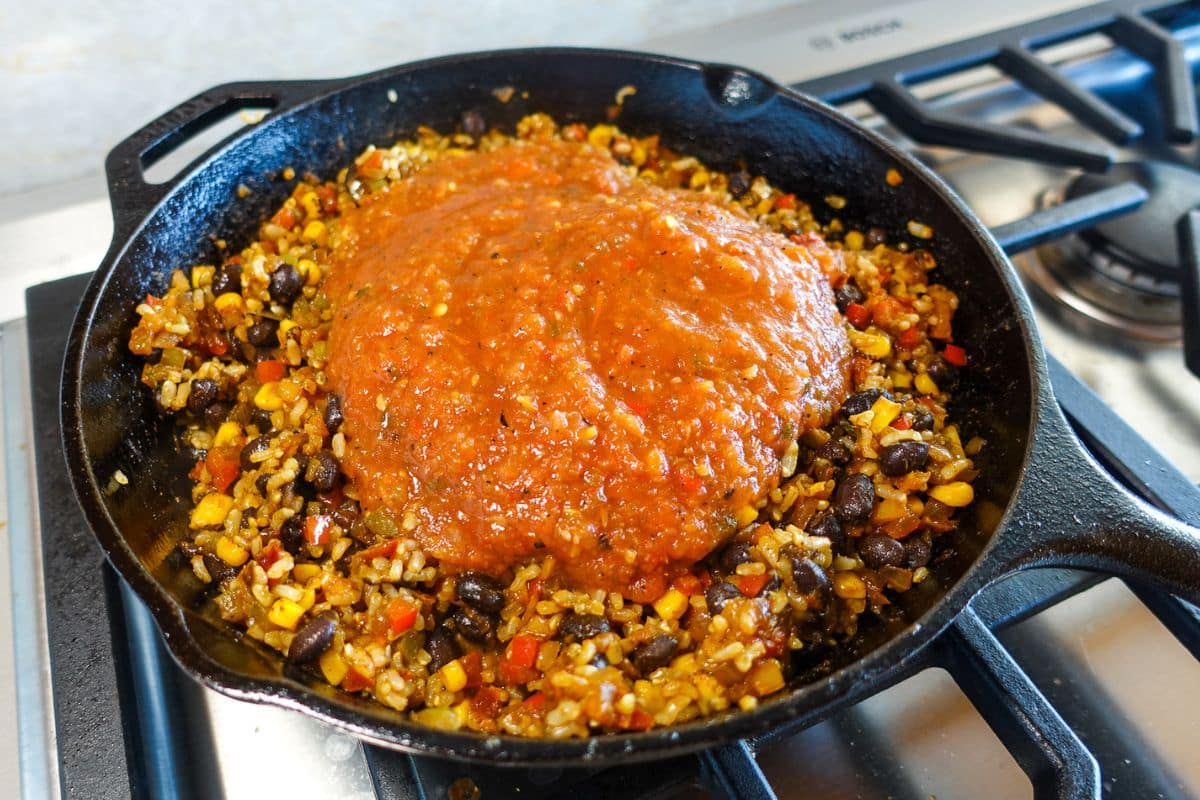 A cast iron skillet on a stove contains rice, black beans, corn, and diced vegetables topped with salsa.