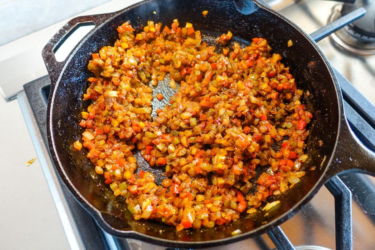 Diced vegetables, including bell peppers and onions with spices, sautéed in a cast iron skillet on a stovetop.