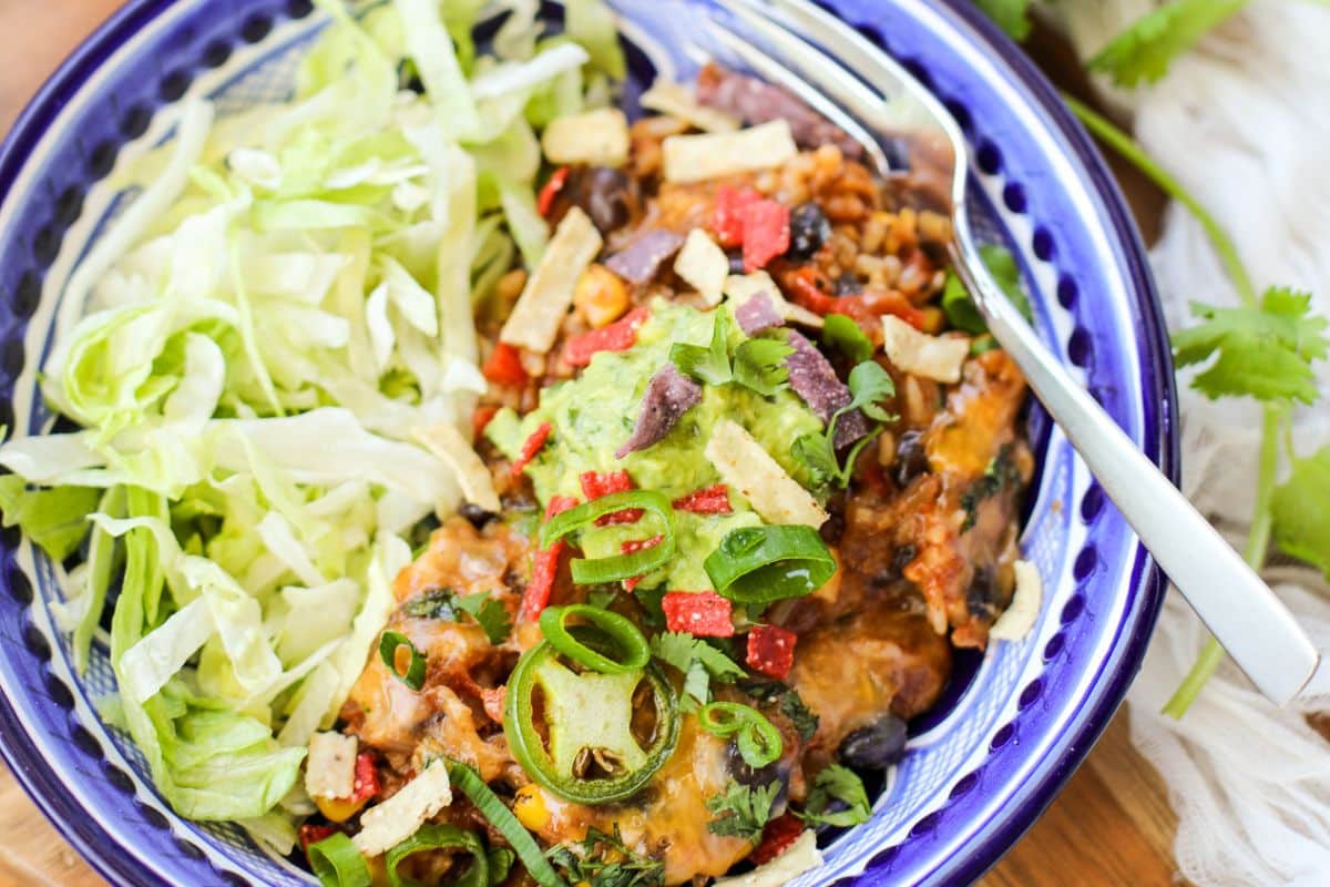 A colorful bowl of Mexican-inspired casserole topped with sliced jalapeños, guacamole, green onions, and tortilla strips, served with shredded lettuce on the side.