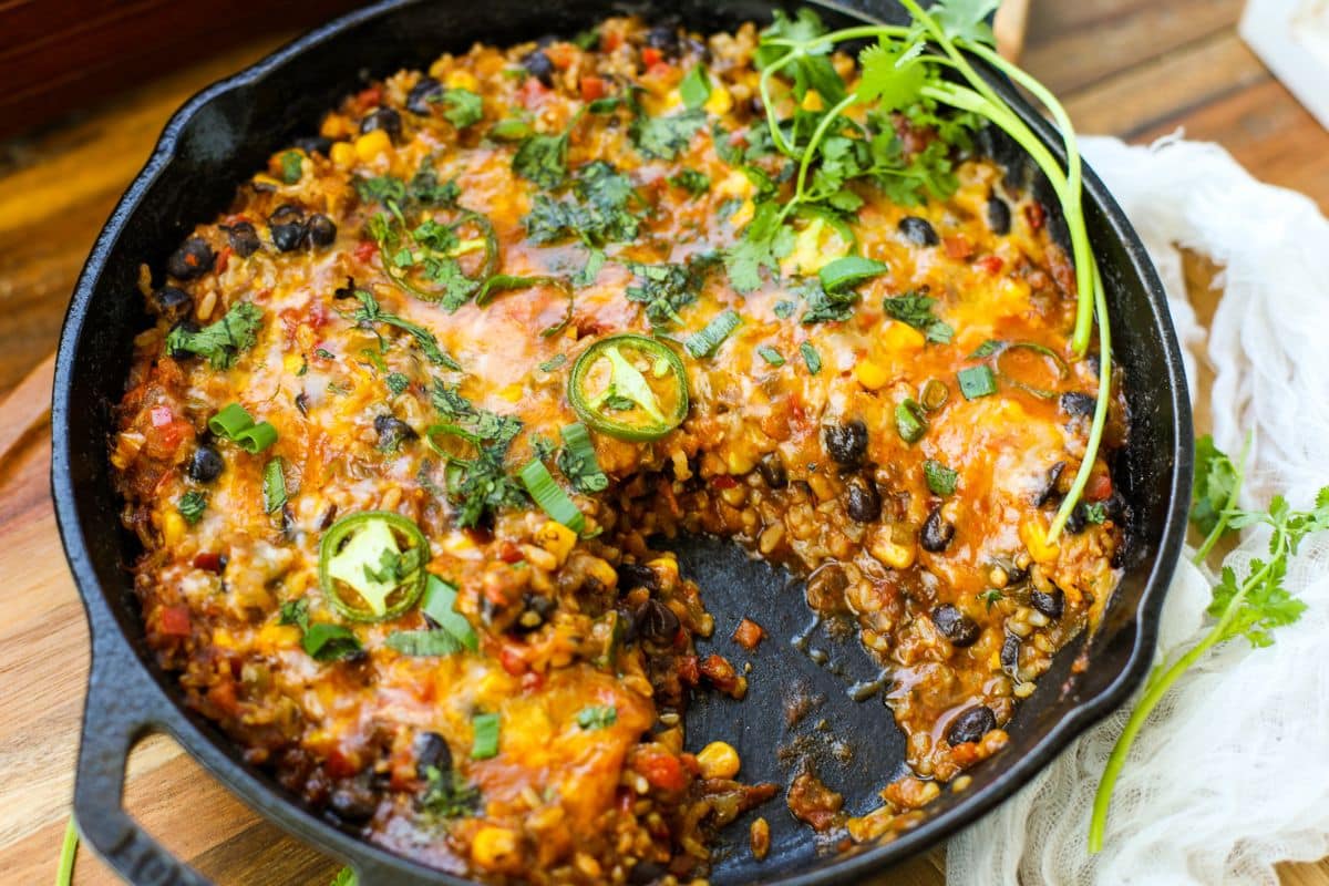 A Vegetarian Burrito Skillet filled with rice, black beans, corn, salsa, melted cheese, spices, and jalapeño slices, with a portion scooped out.