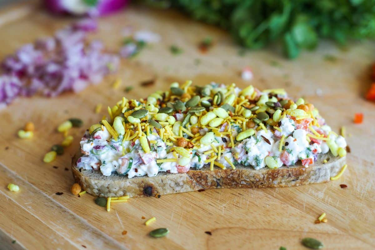 A sourdough toast topped with a creamy vegetable mixture, bhel mix, and pumpkin seeds on a wooden surface with chopped vegetables in the background.
