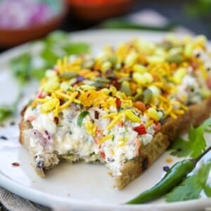 An Indian-inspired cottage cheese toast on sourdough bread topped with veggie cottage cheese, bhel mix, pumpkin seeds, and spices, garnished with cilantro leaves on a white plate.