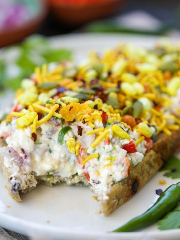 A slice of bread topped with cottage cheese, veggies, bhel mix, pumpkin seeds, and herbs, with a bite taken out. Green chilies and parsley garnish the plate.