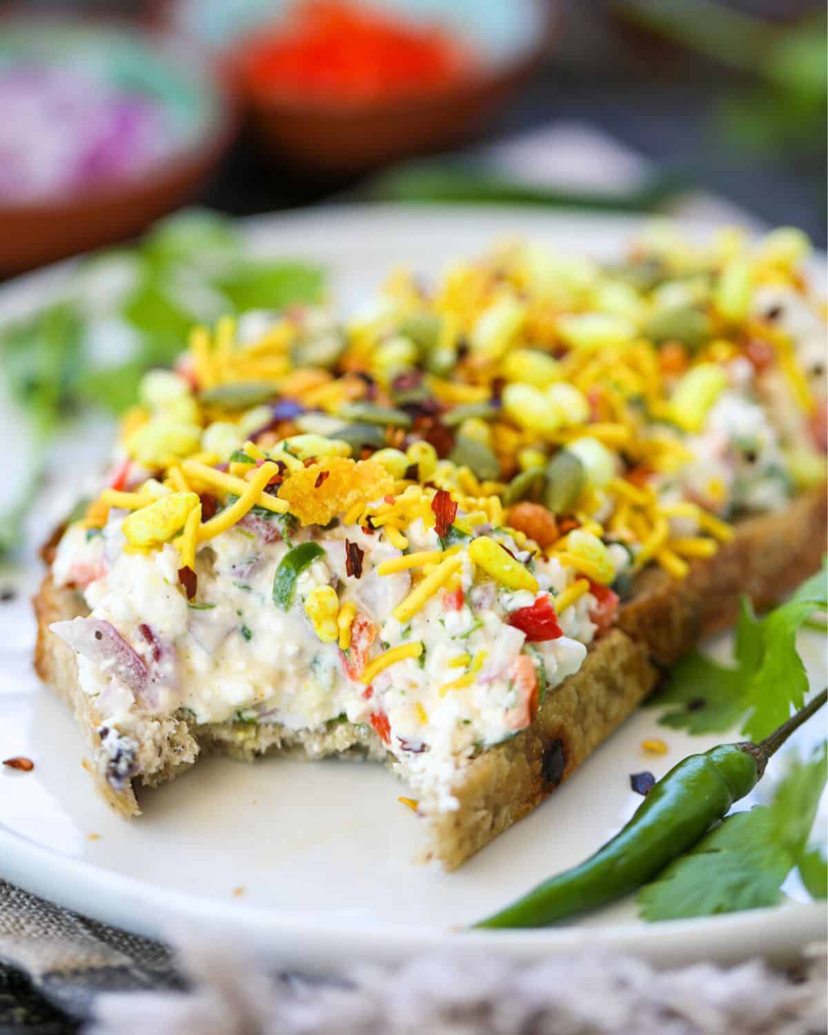 Cottage Cheese Toast on Sourdough (Indian-inspired) contains a slice of bread topped with cottage cheese, veggies, bhel mix, pumpkin seeds, and herbs, with a bite taken out. Green chilies and parsley garnish the plate.
