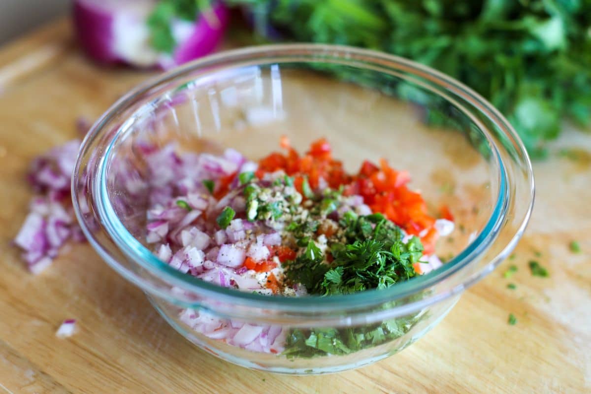 A glass bowl on a wooden surface contains chopped red onions, tomatoes, herbs, and spices. Fresh greens and vegetables are visible in the blurred background.