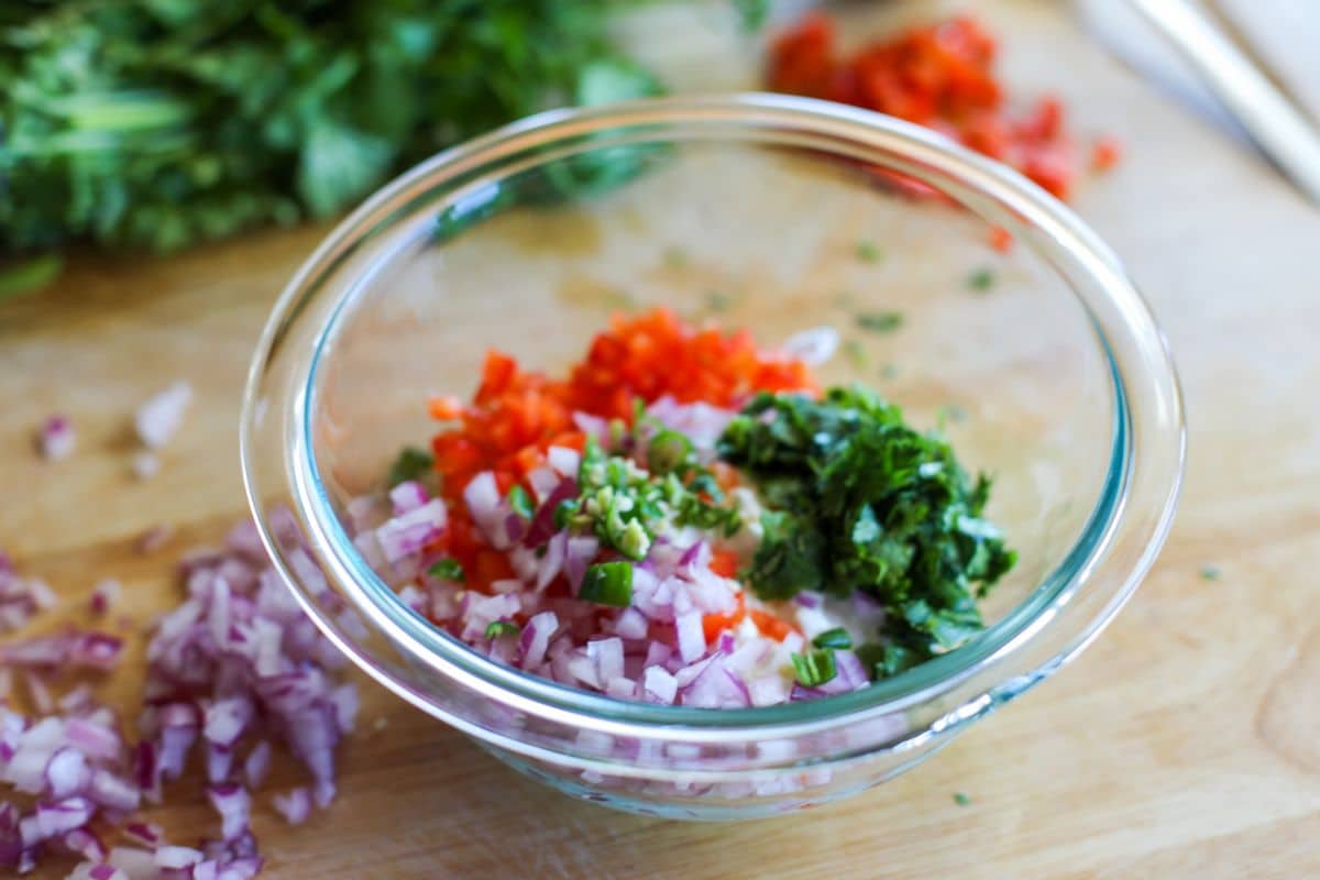 A glass bowl containing chopped red onion, red bell pepper, green chili, and cilantro on a wooden cutting board with more chopped vegetables in the background.