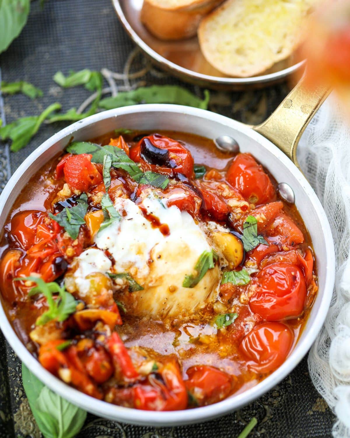 A pan filled with cooked cherry tomatoes, herbs, and a melted burrata cheese center, garnished with fresh basil and served with toasted bread slices in the background.