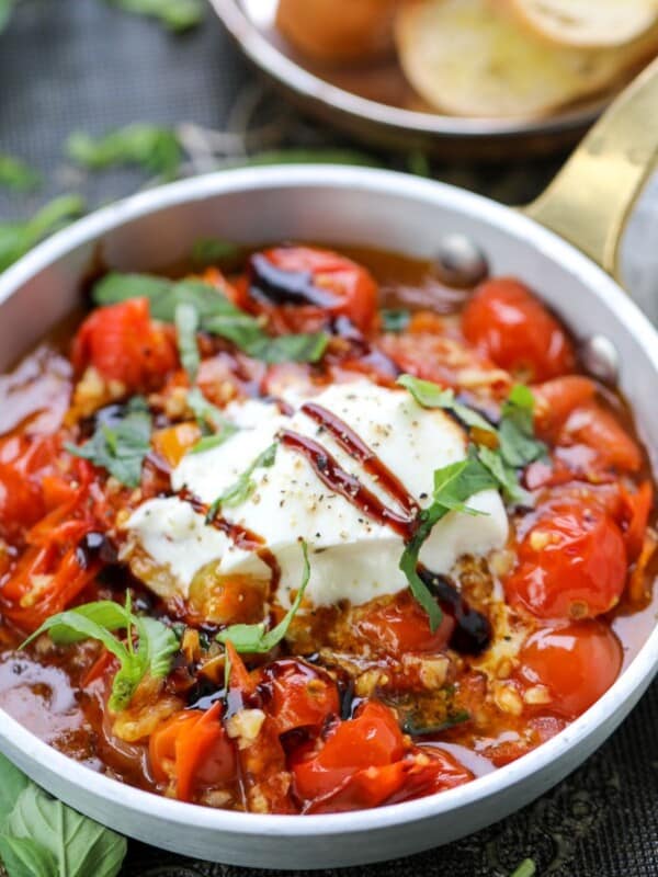 A small pan filled with cooked cherry tomatoes, herbs, and a dollop of soft cheese, garnished with fresh basil and a drizzle of balsamic glaze.