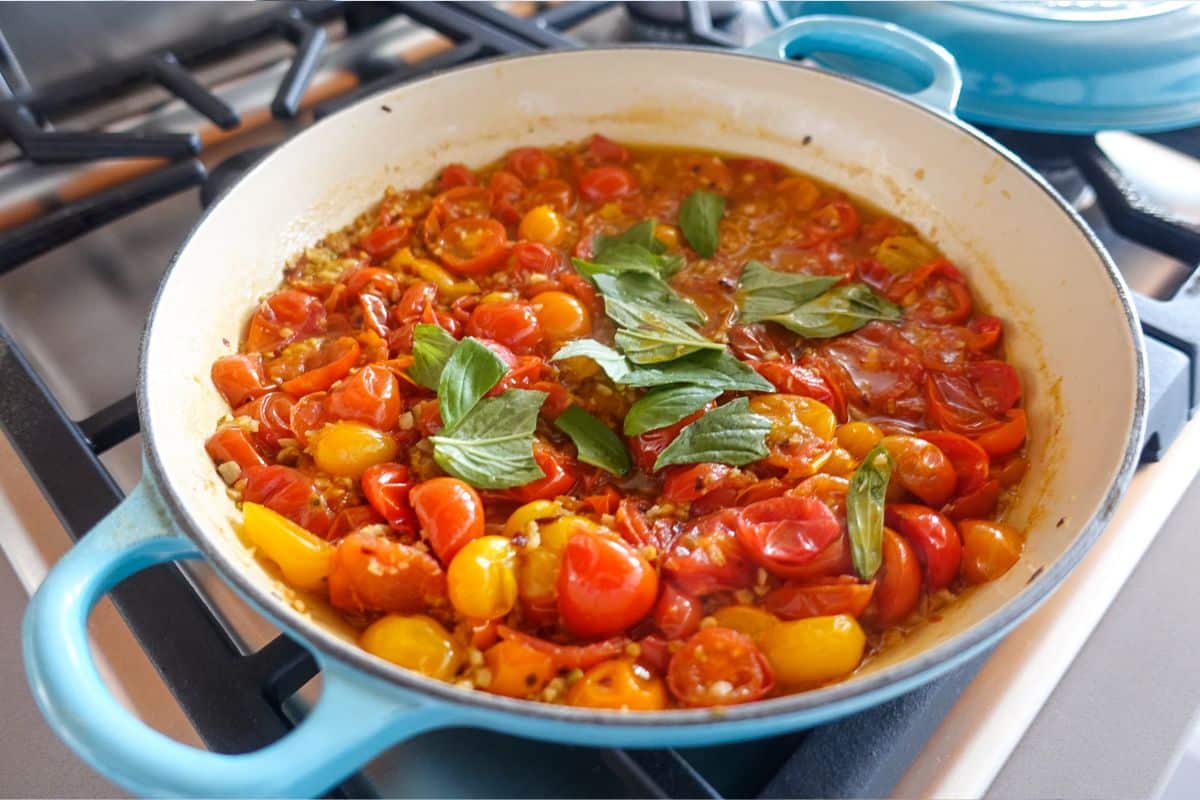 A blue pan on a stove contains a cooked mixture of halved cherry tomatoes and fresh basil leaves in a light sauce.