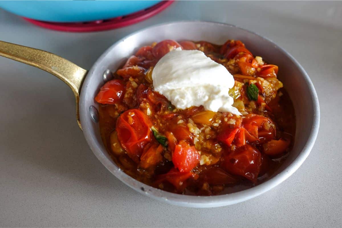 A small pan filled with cooked cherry tomatoes topped with a dollop of burrata cheese, sits on a countertop.
