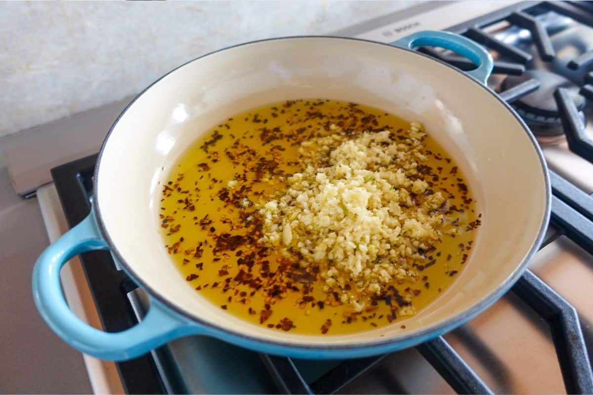 A blue Dutch oven on a stove contains hot oil with minced garlic and red pepper flakes.