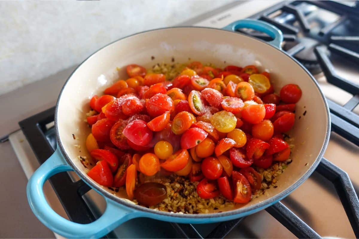 A blue Dutch oven on a stove contains chopped red and yellow cherry tomatoes over saut&eacute;ed garlic.