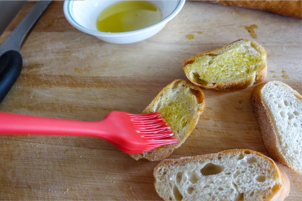 A red brush is spreading olive oil on slices of bread on a wooden cutting board, with a bowl of oil and a knife nearby.