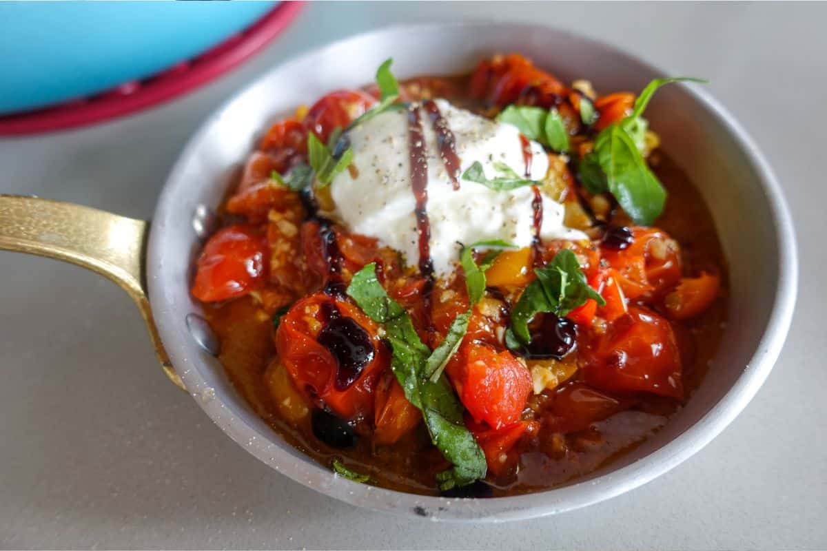 A skillet of roasted cherry tomatoes topped with a ball of burrata cheese, fresh basil, and a drizzle of balsamic glaze.