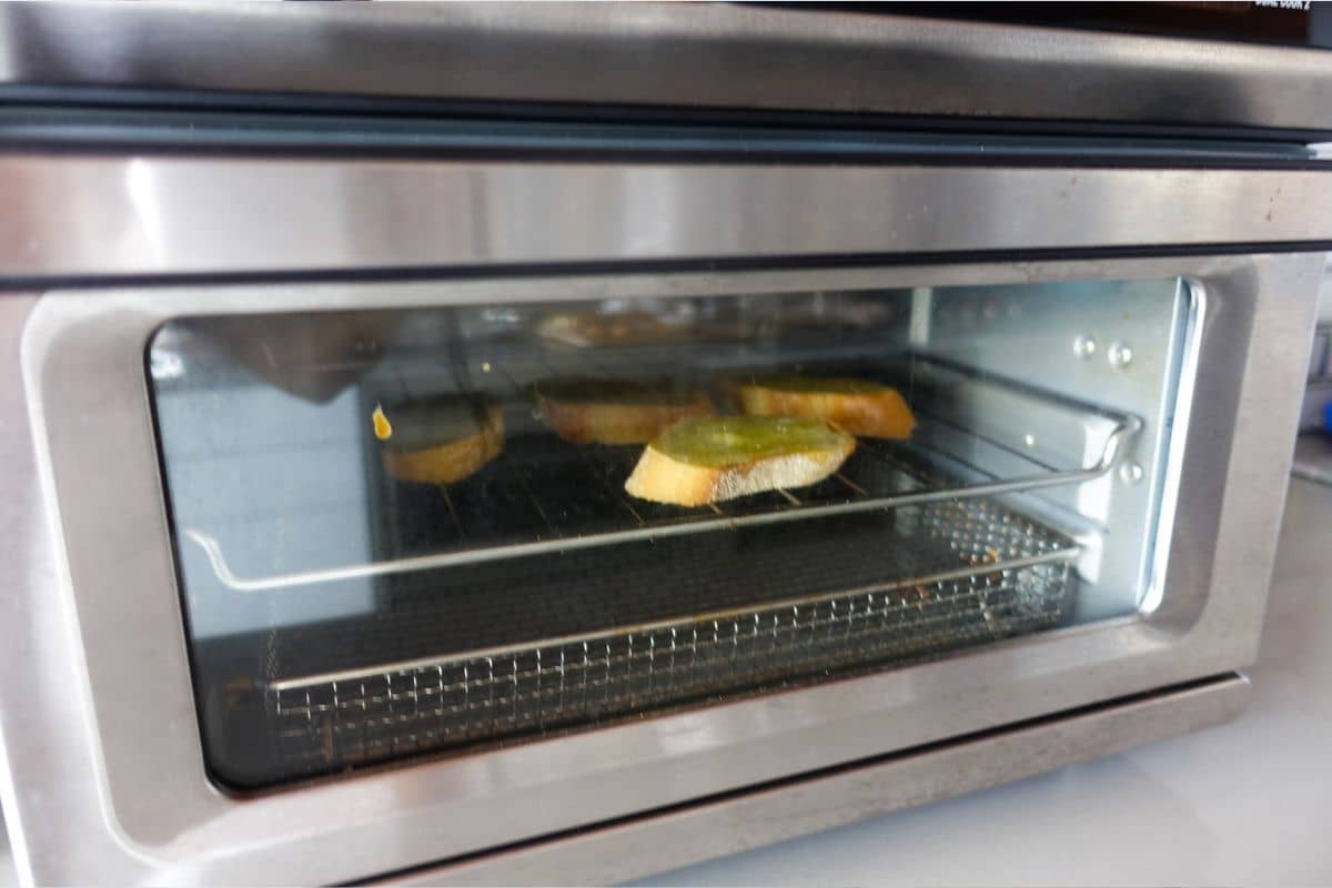 Slices of bread with olive oil are being toasted on a rack inside a stainless steel toaster oven.