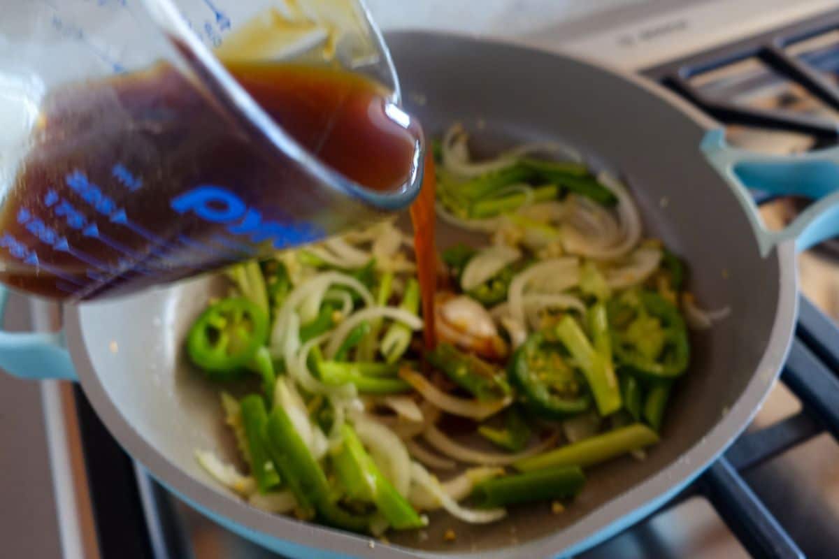 Add Asian sauce from a measuring cup into a pan with veggies on a gas stove.