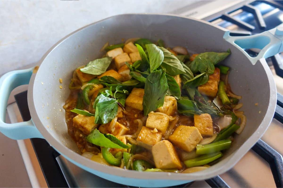 A skillet on a stove contains tofu cubes, green bell peppers, onions, and fresh Thai basil leaves in a brown sauce.