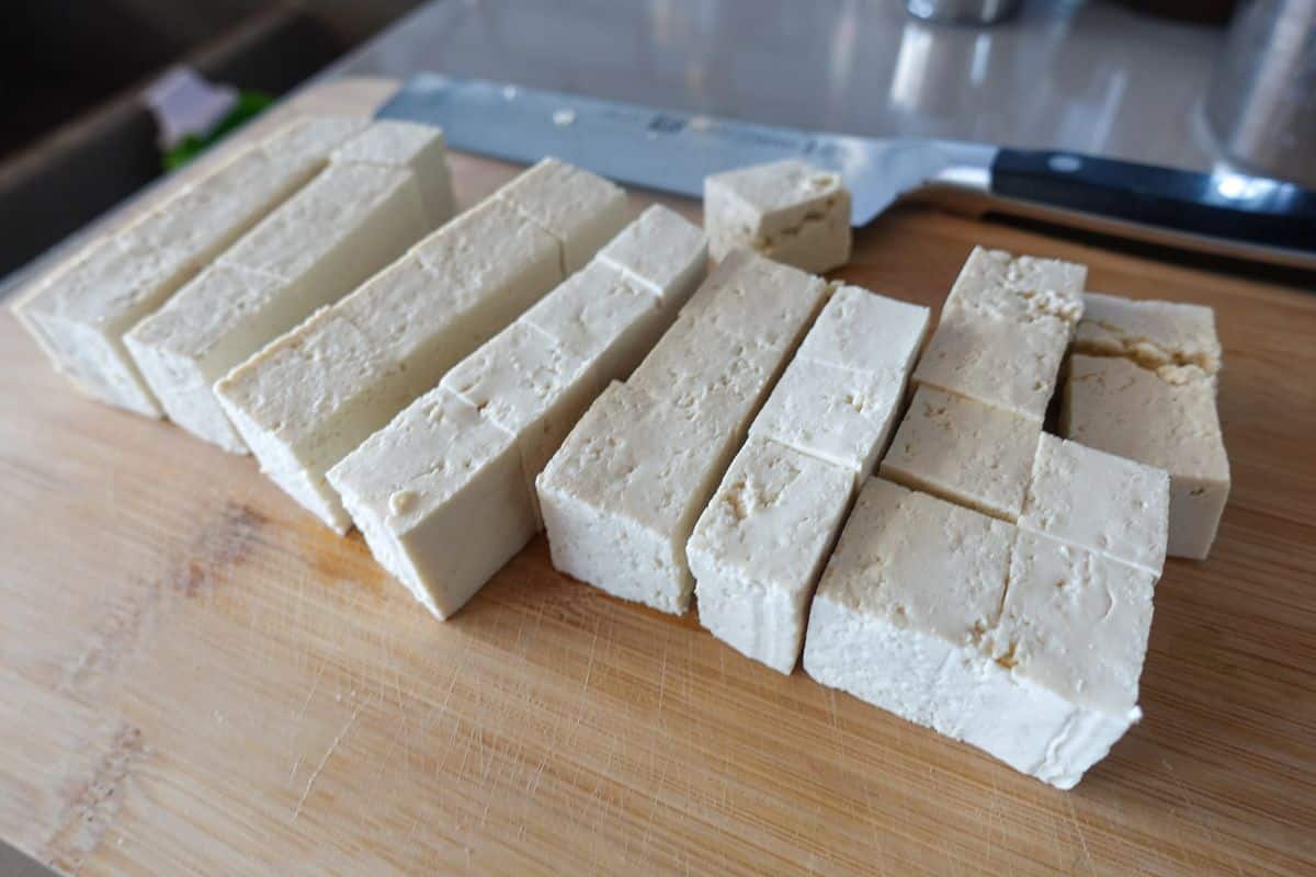 Blocks of firm tofu cut into rectangular pieces on a wooden cutting board with a knife in the background.