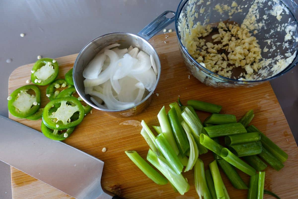 Sliced scallions, onion, and jalapeño, and minced garlic on a wooden cutting board with a knife and measuring cup.