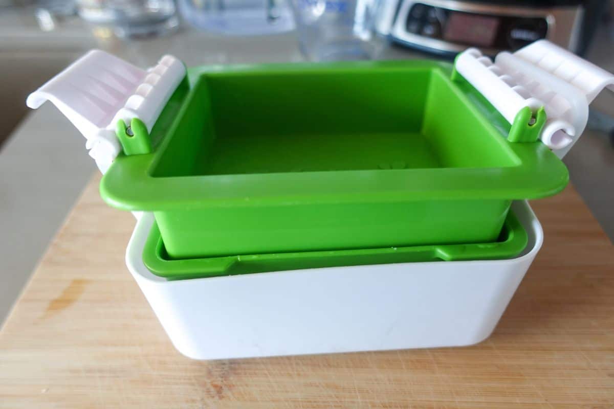 A green and white plastic tofu press sits on a wooden surface, with clamping lids open on both sides.