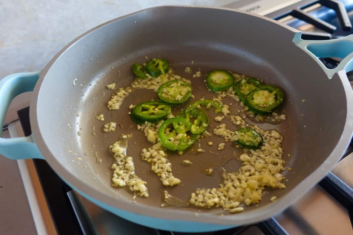 Sliced jalapeños and minced garlic are sautéing in oil in a light blue frying pan on a stovetop.