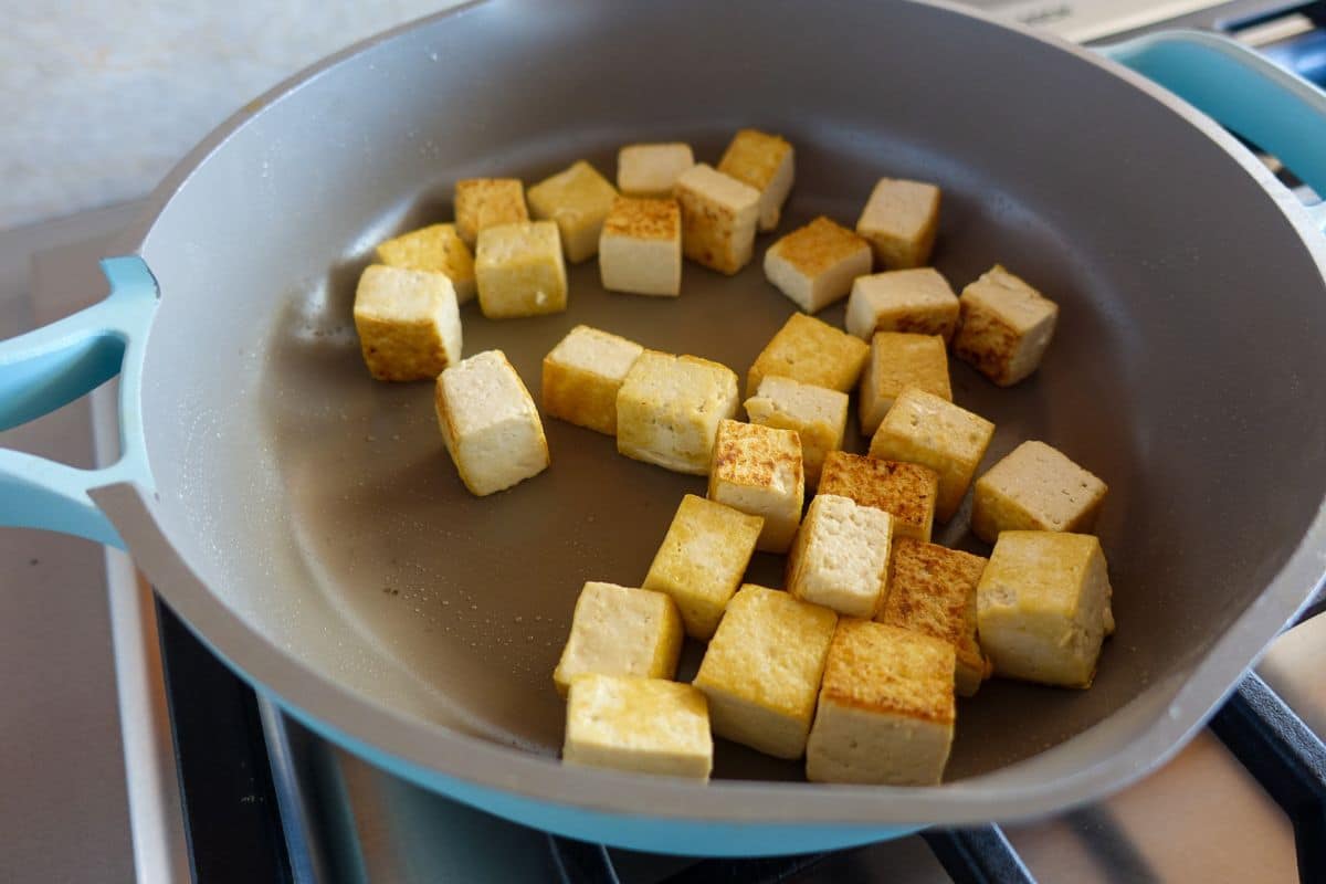 A frying pan on a stove contains cubed pieces of tofu being sautéed, some sides golden brown.