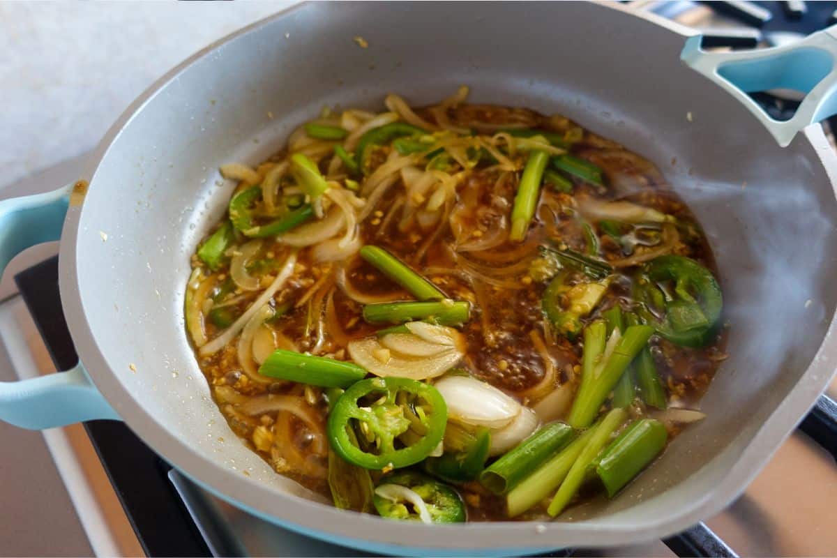 A light blue pan on a stove contains sautéed veggies and Asian sauce that's sizzling.