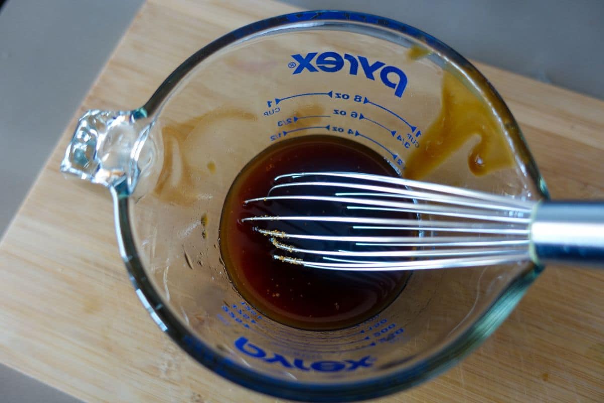 A glass measuring cup with Asian sauces and a metal whisk inside, placed on a wooden surface.