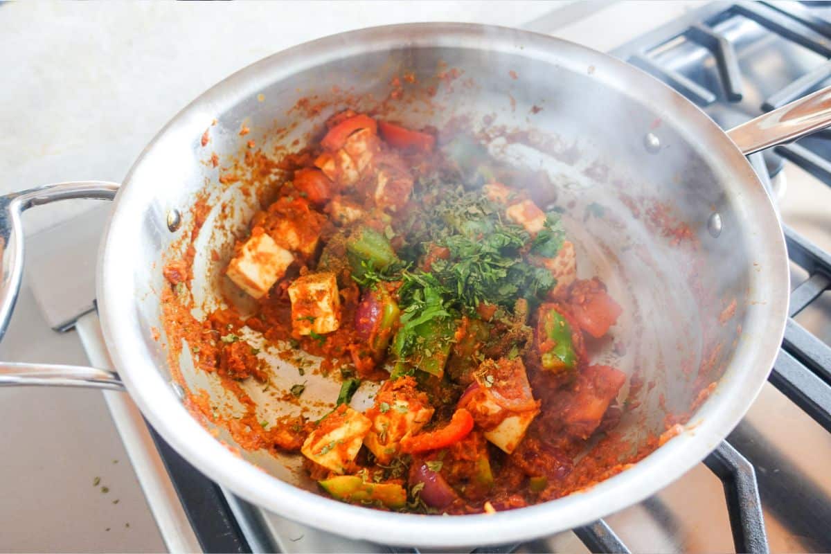 A stainless steel pan on a stove contains a simmering paneer with visible herbs, bell peppers, and sauce.