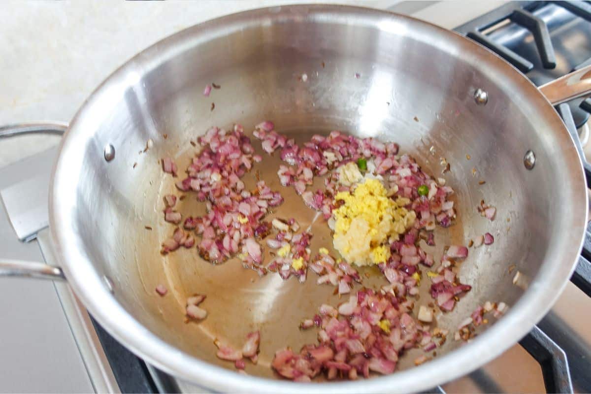 Diced red onions and minced garlic, ginger, and green chilies saut&eacute;ing in a stainless steel pan on a stovetop.