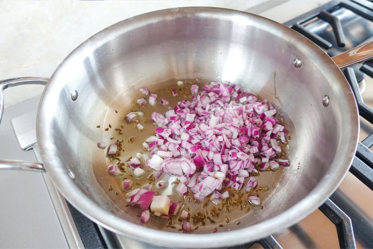 Chopped red onions and cumin seeds saut&eacute;ing in oil in a stainless steel pan on a gas stove.