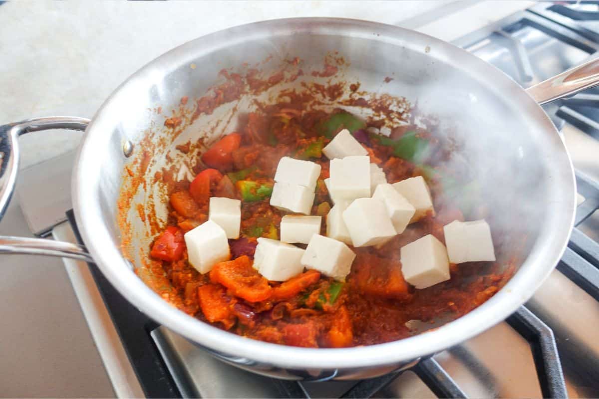 A stainless steel pan on a stove contains a tomato-based sauce with vegetables and cubes of paneer. Steam rises from the pan.
