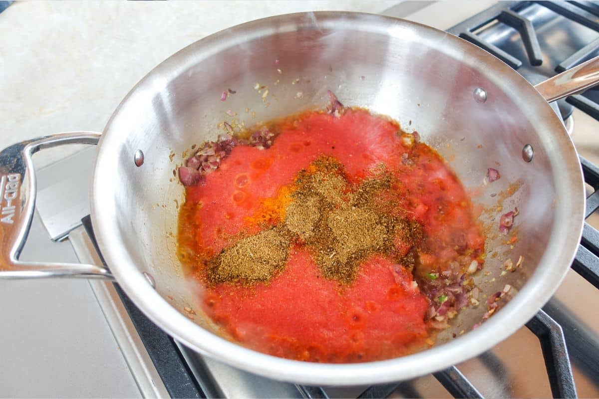 A stainless steel pan on a stove contains saut&eacute;ed onions, tomato puree, and a mix of ground spices that have just been added but not yet stirred.