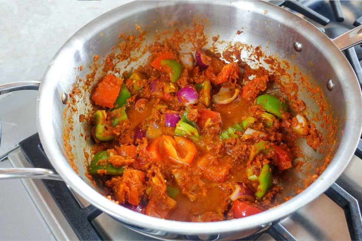 A stainless steel pan on a stove contains a colorful mixture of chopped tomatoes, bell peppers, onions, and spices being saut&eacute;ed.
