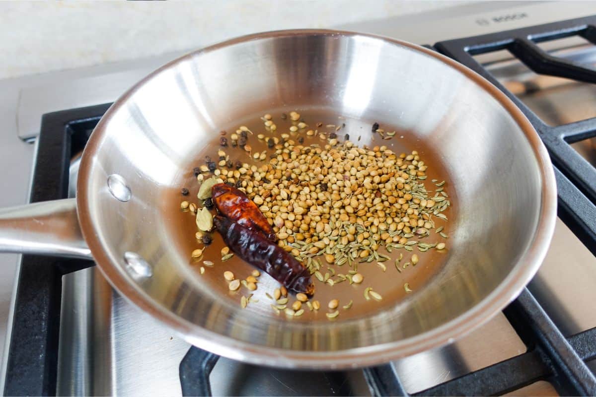 A stainless steel pan on a stove with whole spices including dried red chili, coriander seeds, fennel seeds, and peppercorns being dry roasted.