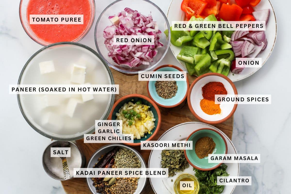 A variety of labeled ingredients for making kadai paneer, including paneer, tomato puree, onions, bell peppers, spices, ginger, garlic, green chilies, oil, and cilantro.
