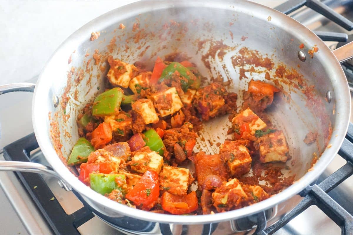 A stainless steel pan on a stovetop contains cooked paneer cubes, bell peppers, and tomatoes in a spiced tomato sauce. Kadai Paneer is ready.