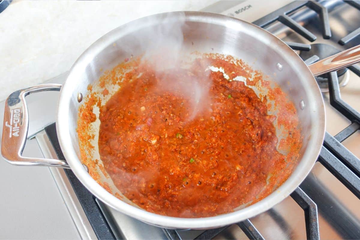 A stainless steel pan with simmering red tomato puree on a stove, with visible steam rising from the sauce.